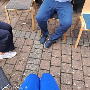 mehre Menschen, von denen man nur die Beine sieht, sitzen auf Stühlen im Hof